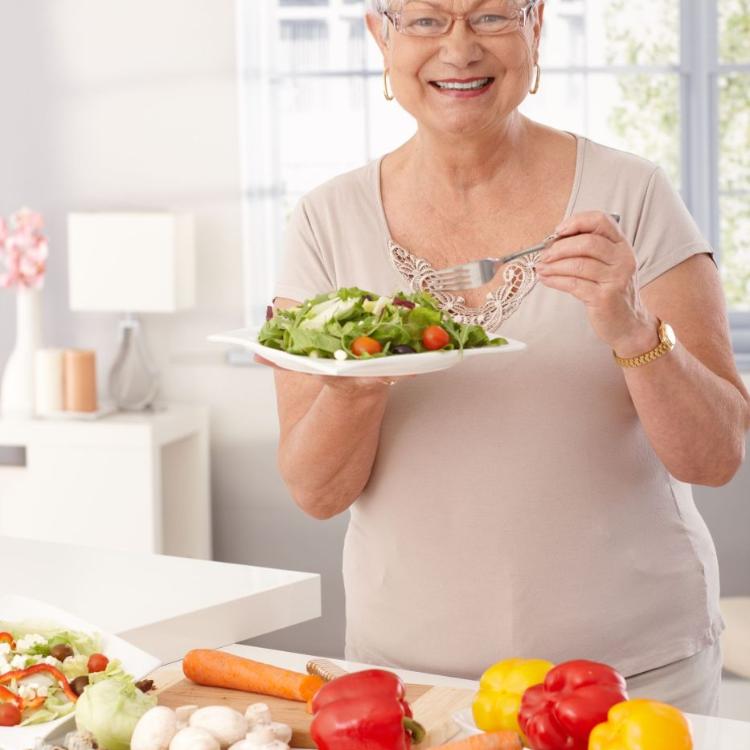 26224867 - modern grandmother eating fresh green salad and vegetables in kitchen, smiling happy, looking at camera.