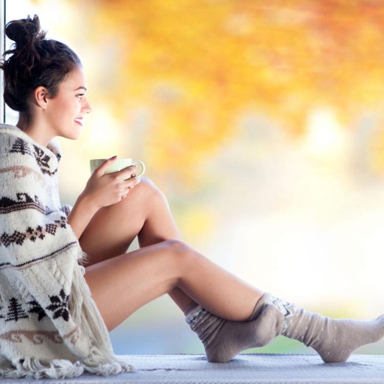 45215465 - young beautiful brunette woman with cup of coffee wearing knitted nordic print poncho sitting home by the window. blurred garden fall background.